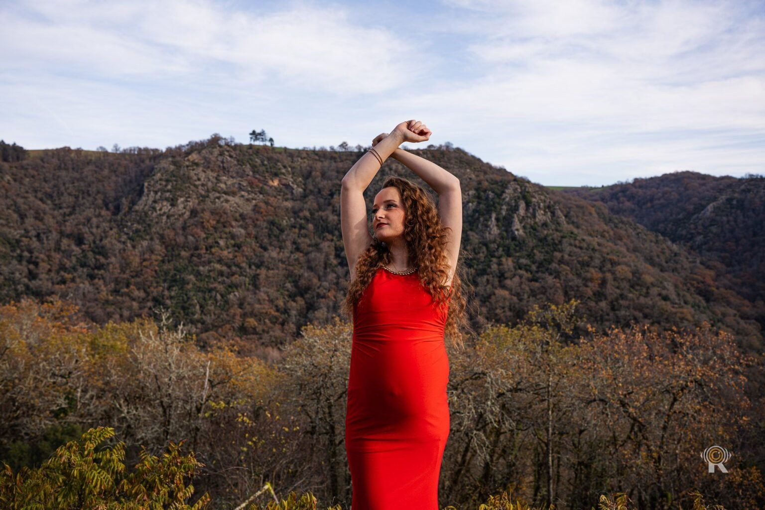 Une femme enceinte en robe rouge, bras levés, dans la montagne.