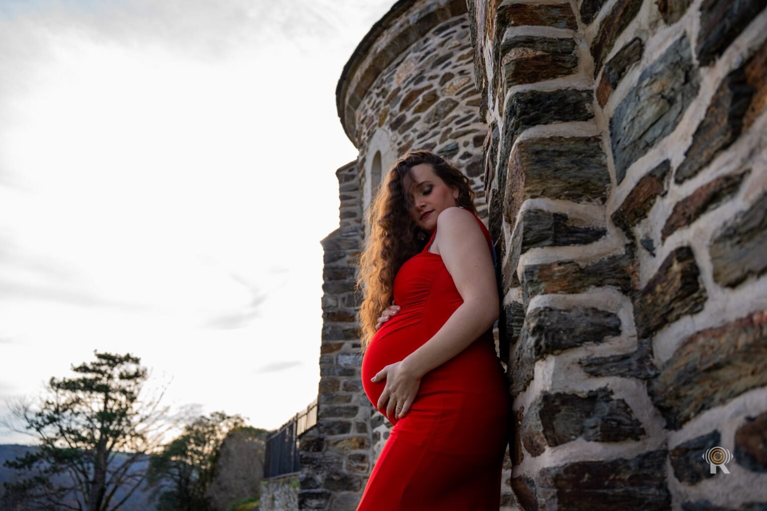 Une femme enceinte en robe rouge, appuyée contre une tour en pierre.