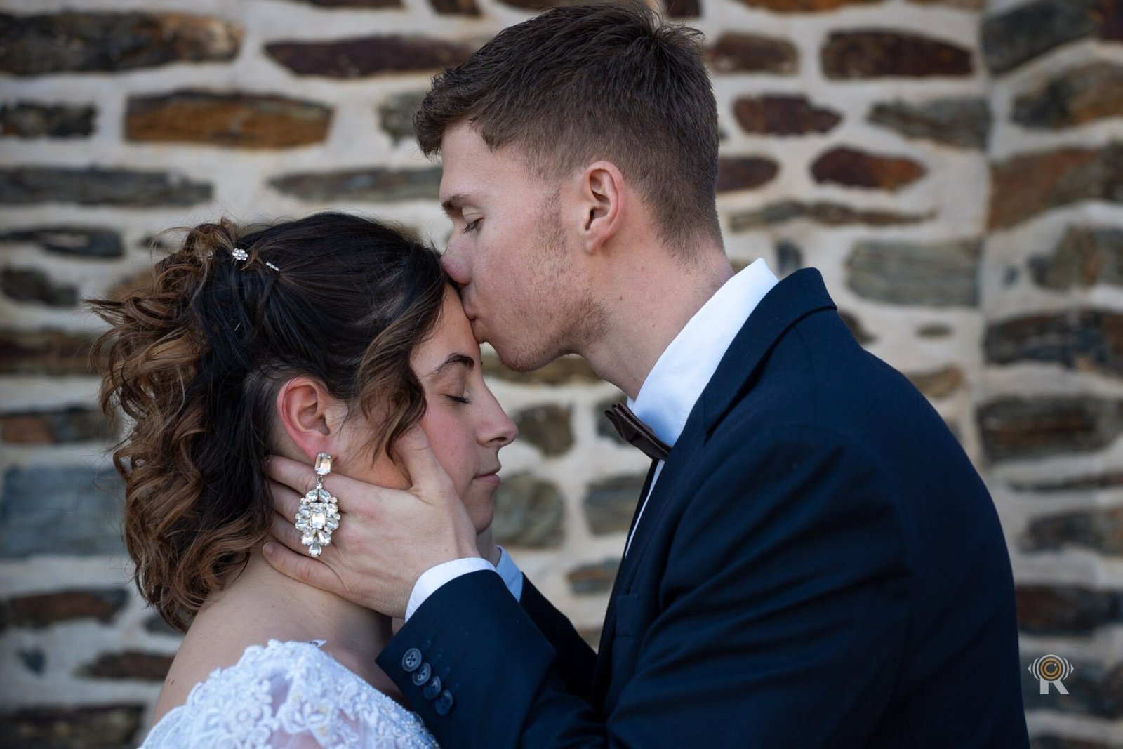 Le marié embrasse le front de la mariée devant un mur de pierre, mariage à Toulouse.