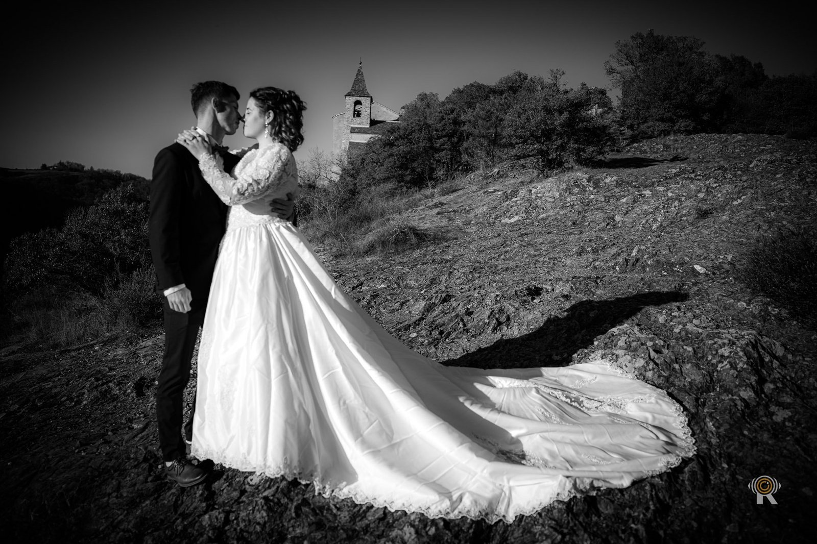 Les mariés sur une colline devant une église, robe déployée, noir et blanc.