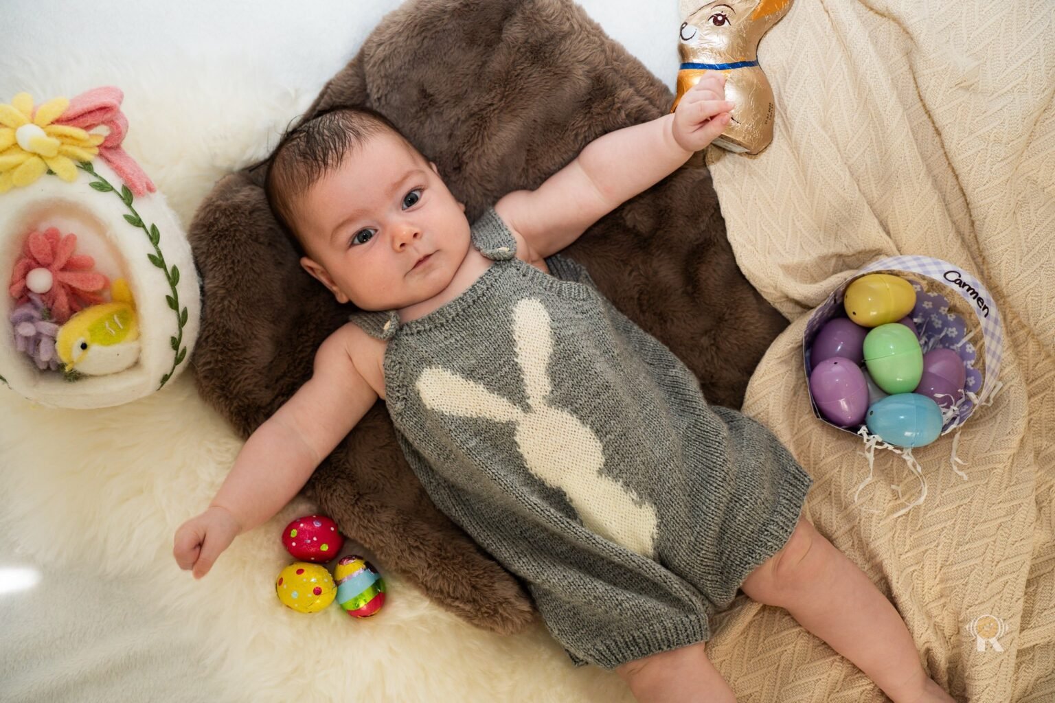 Un bébé allongé, tenant un lapin en chocolat, entouré d'œufs de Pâques.