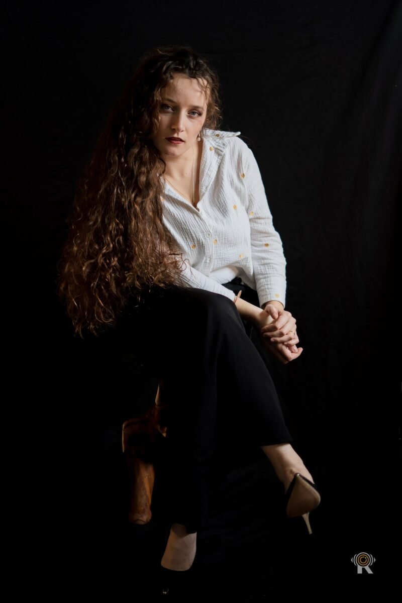 Jennifer Igreja assise sur un tabouret, jambes croisées, mains jointes sur le genou, regard vers l’objectif