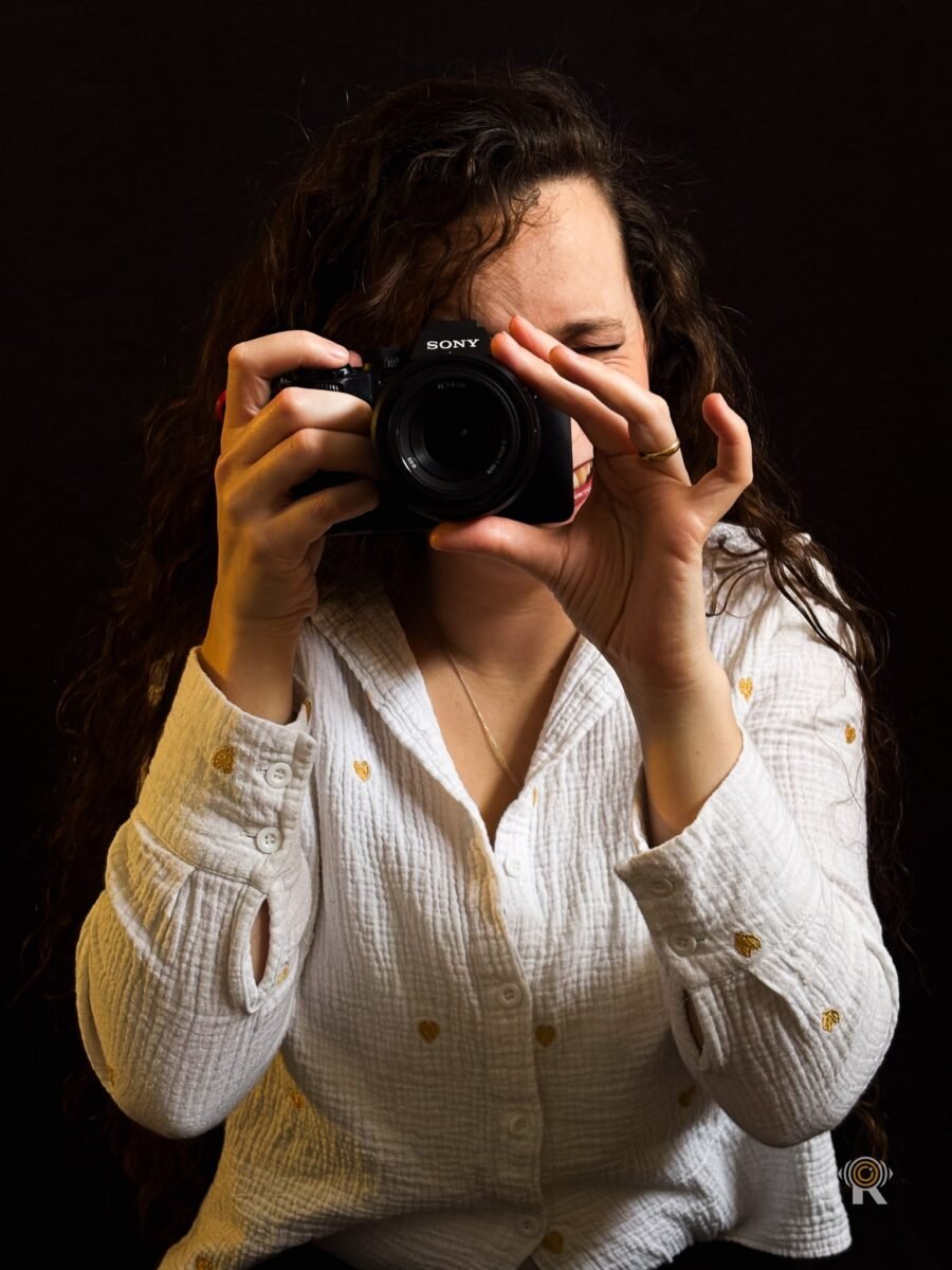 Jennifer Igreja rit derrière un boîtier Sony levé à l’œil, chemise blanche à petits motifs dorés