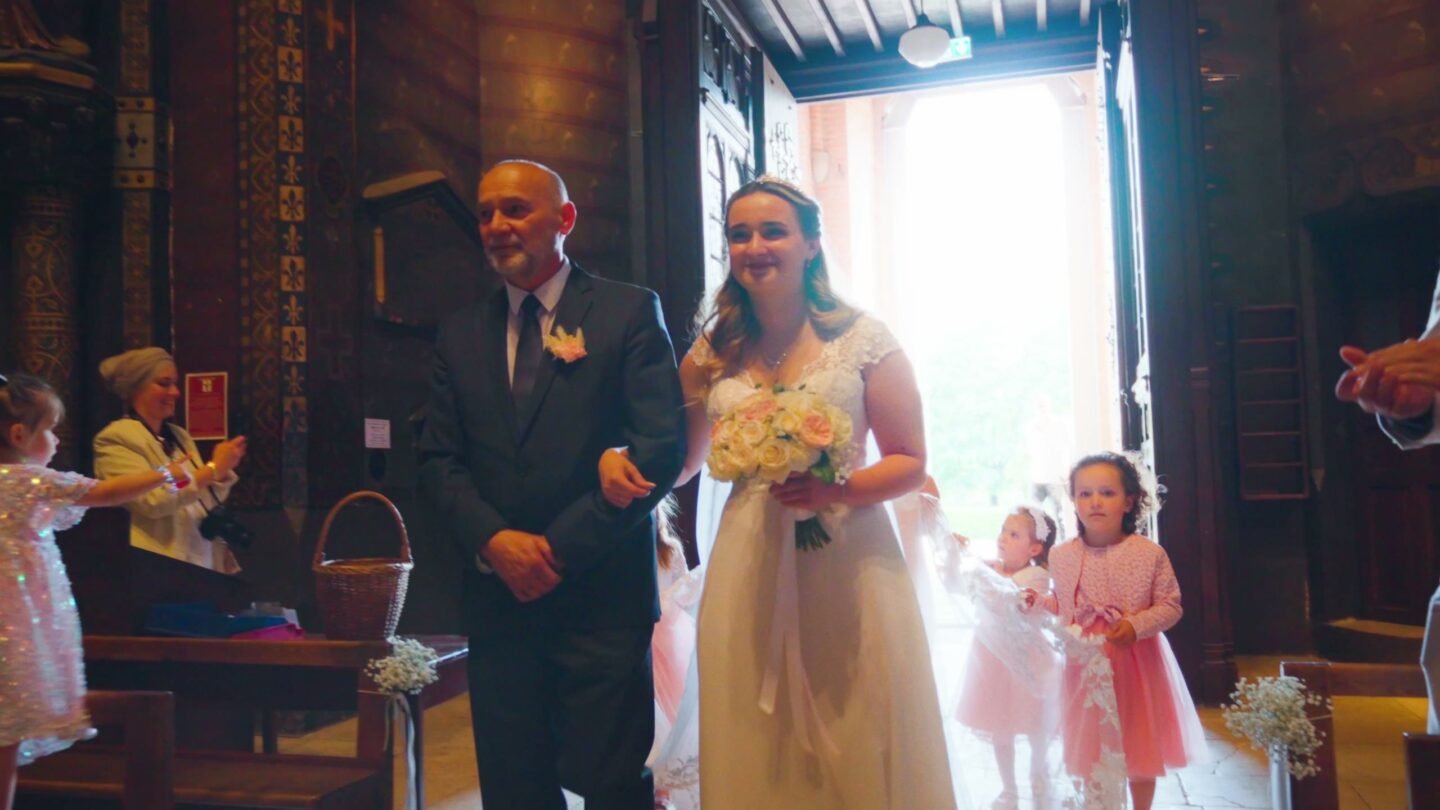 Dans l’église, la mariée avance au bras de son père, à contre-jour, suivie de deux petites filles en robe rose.