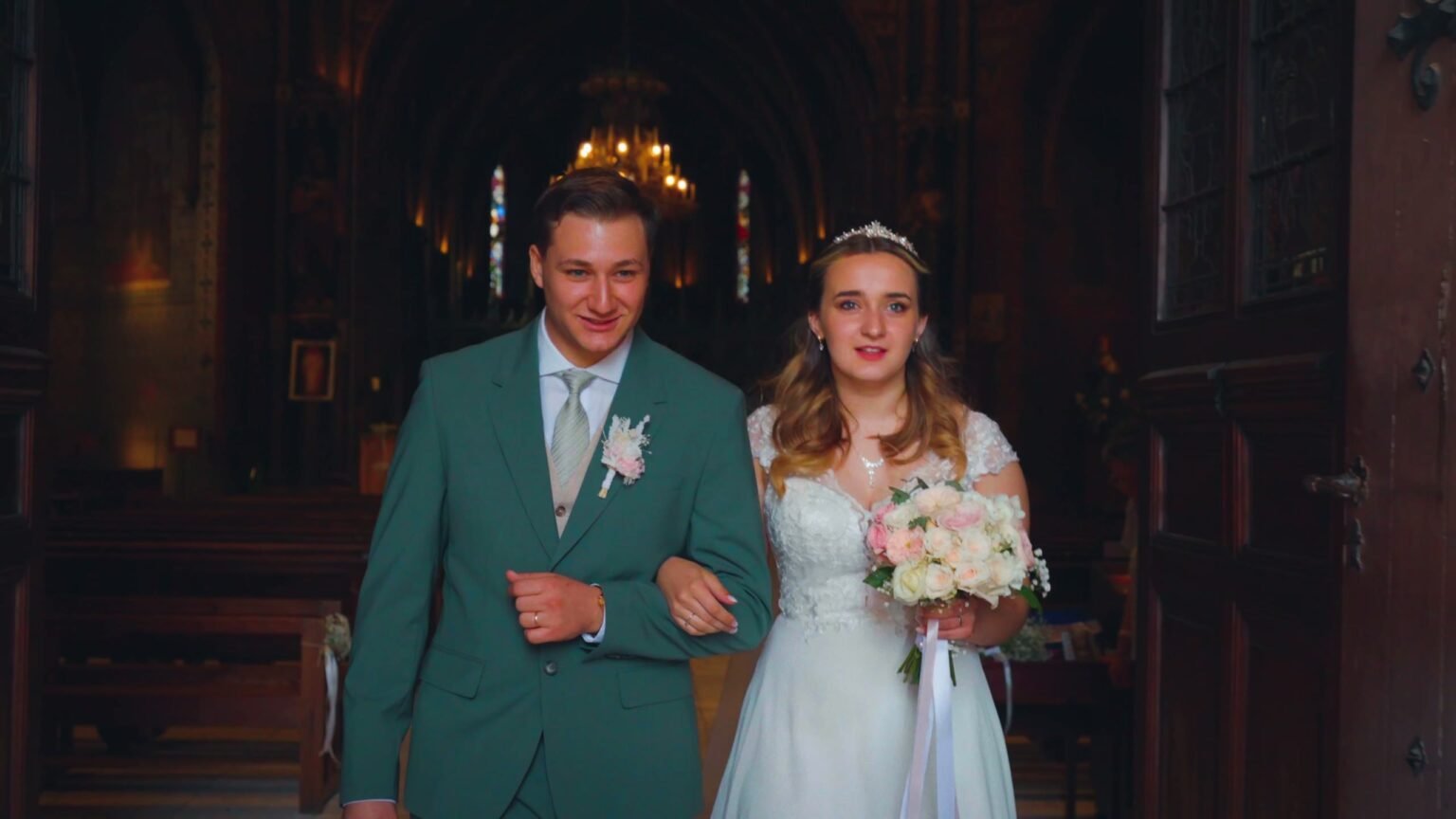 Les mariés sortent de l’église bras dessus bras dessous, lui en costume vert, elle avec son bouquet de roses.