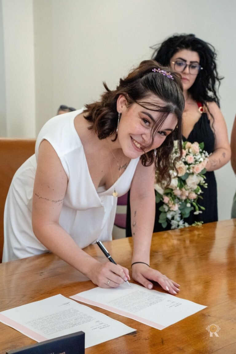 À la mairie, la mariée sourit en signant le registre, son témoin derrière elle avec le bouquet.