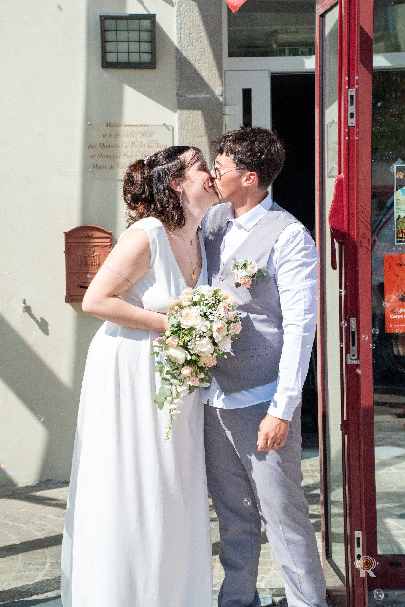 Les mariés s’embrassent devant la porte rouge de la mairie, elle tient son bouquet.