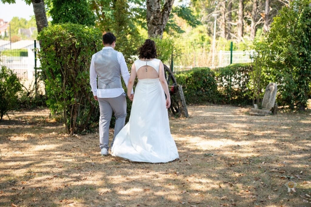 Les mariés s’éloignent main dans la main dans le jardin, robe blanche de dos