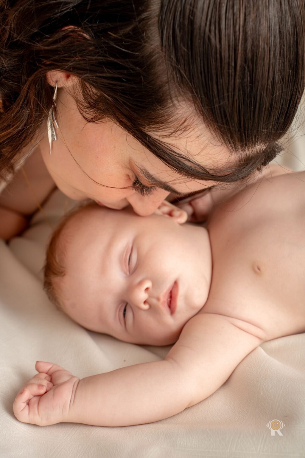 Mère embrassant son bébé endormi, la joue contre sa tête, sur un drap blanc dans une lumière douce