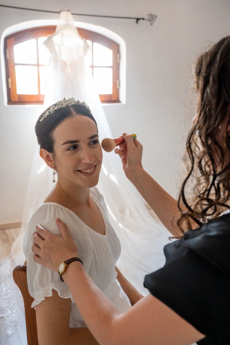 La mariée sourit pendant qu’on la maquille, sa robe en arrière-plan.