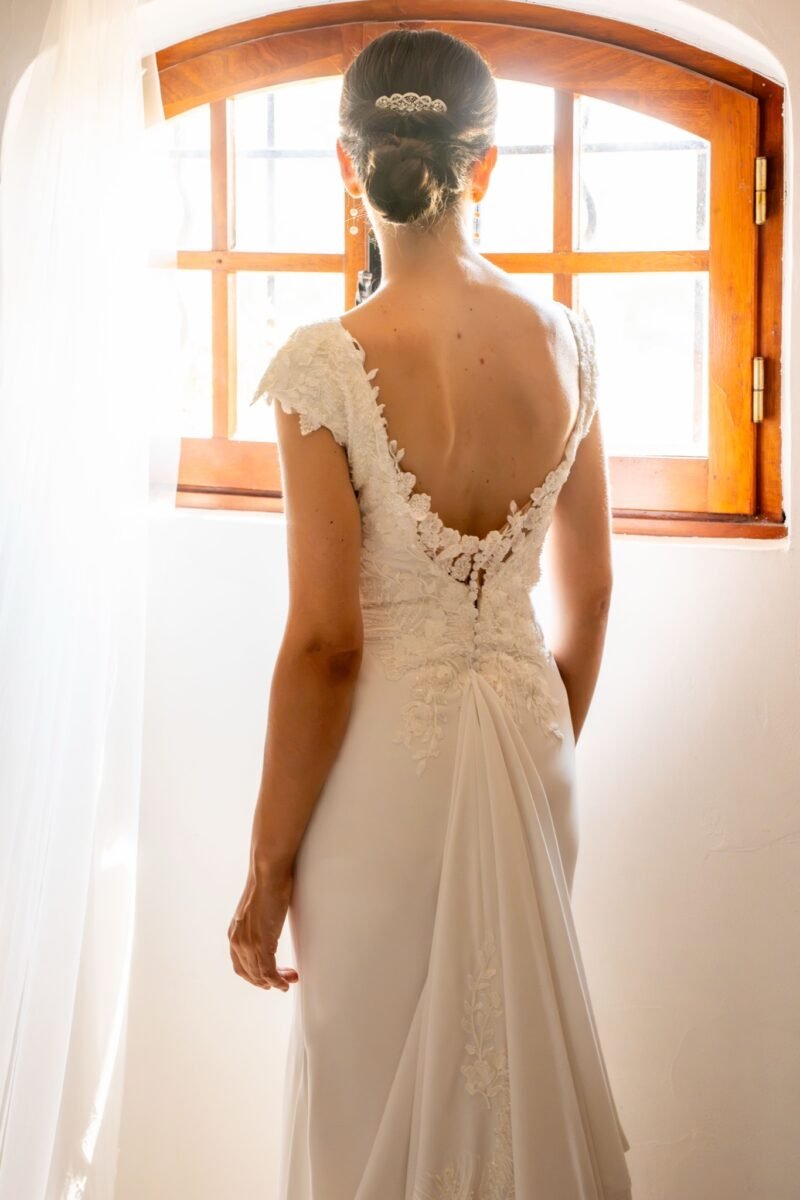 La mariée de dos devant la fenêtre, on voit la dentelle de sa robe.