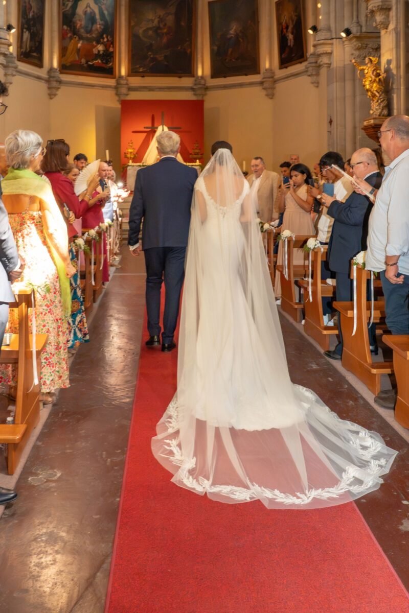 La mariée de dos dans la nef, son long voile étalé sur le tapis rouge.