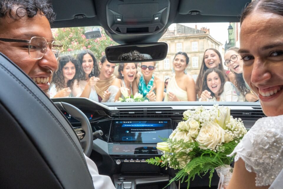 Des proches crient devant le pare-brise, les mariés à l’intérieur de la voiture.